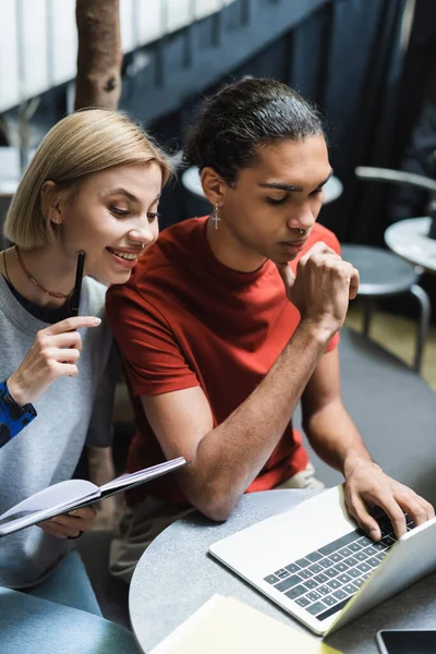 Freelancer positivo sosteniendo portátil cerca de amigo afroamericano y dispositivos en la cafetería - foto de stock