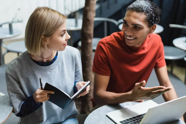 Positive african american freelancer pointing at laptop near friend with notebook in cafe — Stock Photo