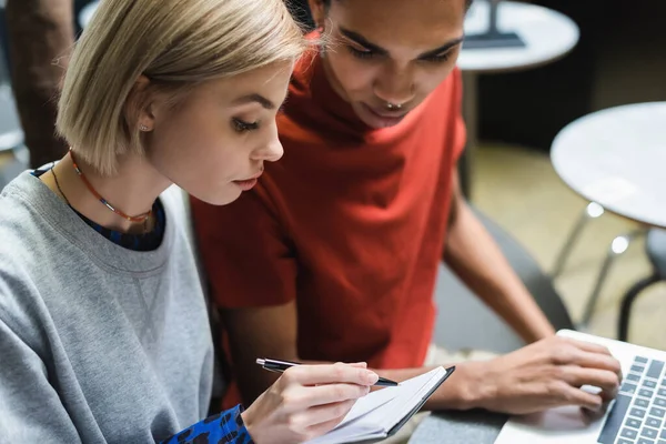 Young freelancer writing on notebook near african american friend and laptop in cafe — Stock Photo