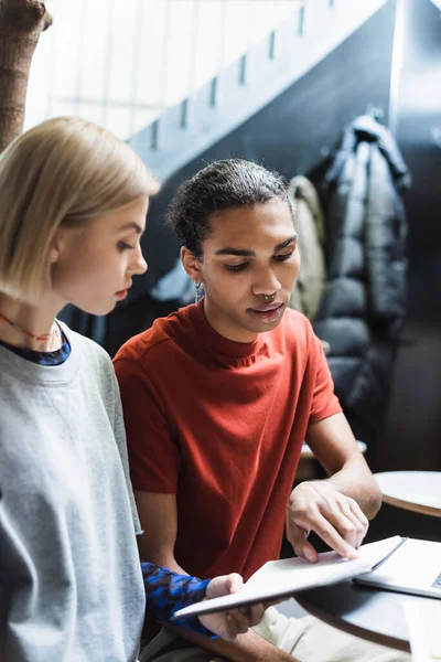 African american freelancer pointing at notebook near friend and blurred laptop in cafe — Stock Photo