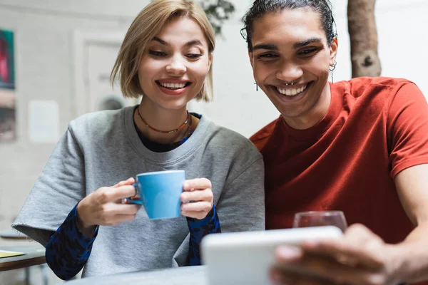 Sonriente pareja interracial con café mirando el teléfono inteligente borroso en la cafetería - foto de stock