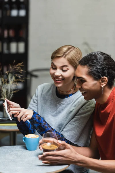 Alegre pareja multiétnica utilizando el teléfono inteligente cerca del café en la cafetería - foto de stock