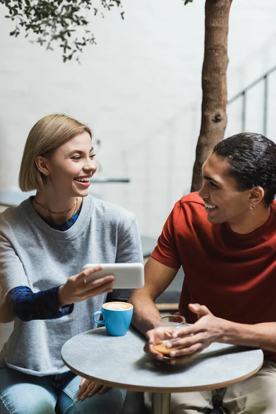 Lächelnder afrikanisch-amerikanischer Mann schaut Freundin mit Smartphone in der Nähe von Kaffee im Café an — Stockfoto