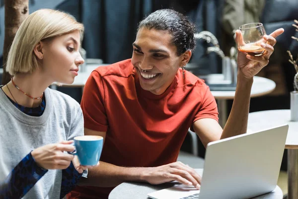 Positive african american freelancer holding coffee near laptop and friend in cafe — Stock Photo