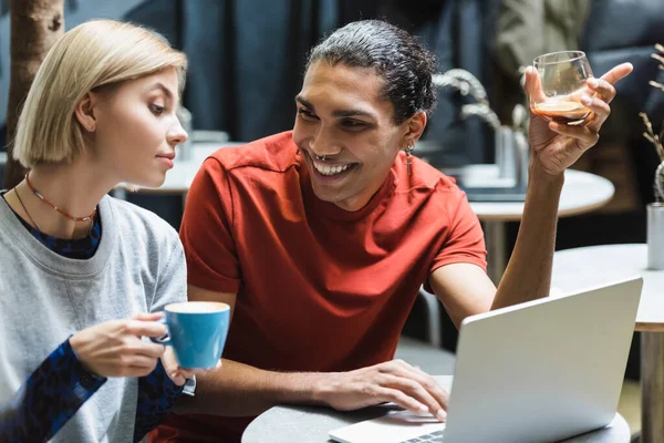 Positivo afroamericano freelancer celebración de café cerca de la computadora portátil y amigo en la cafetería - foto de stock