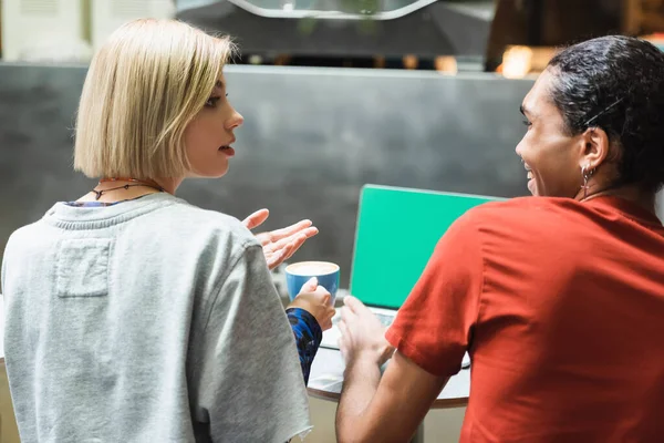 Side view of multiethnic freelancers with coffee talking near laptop with green screen in cafe — Stock Photo