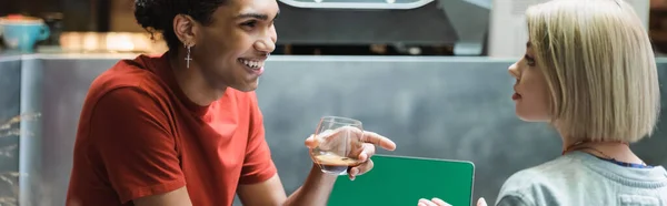 Freelancer afro-americano sorridente segurando café perto de amigo e laptop no café, banner — Fotografia de Stock