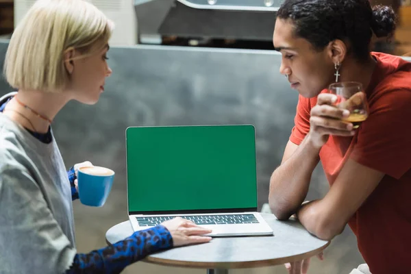 Side view of multiethnic freelancers holding coffee and using laptop with green screen in cafe — Stock Photo