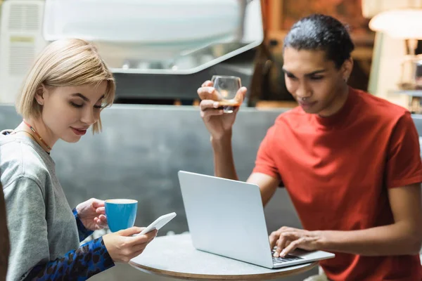 Mujer joven con café usando teléfono inteligente cerca de amigo afroamericano con computadora portátil en la cafetería - foto de stock