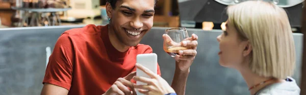 Mujer sonriente sosteniendo el teléfono celular cerca de novio afroamericano con café en la cafetería, pancarta - foto de stock