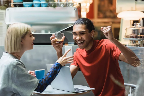 Excited african american freelancer holding coffee near girlfriend and devices in cafe — Stock Photo