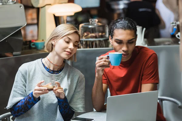 Jóvenes freelancers multiétnicos sosteniendo café cerca de aparatos en la cafetería - foto de stock