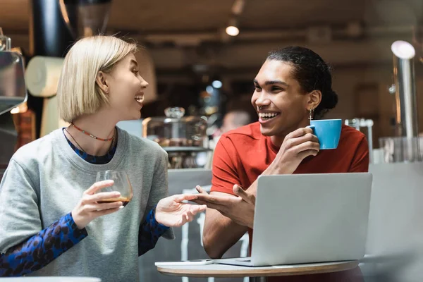 Smiling interracial freelancers talking and holding coffee near devices in cafe — Stock Photo