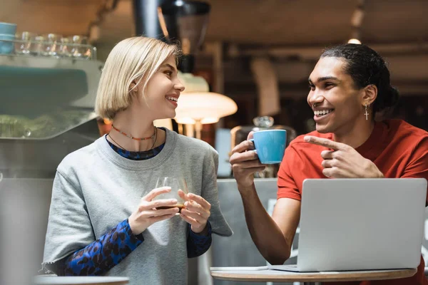 Freelancer afro-americano positivo segurando café e apontando para o amigo perto de dispositivos no café — Fotografia de Stock