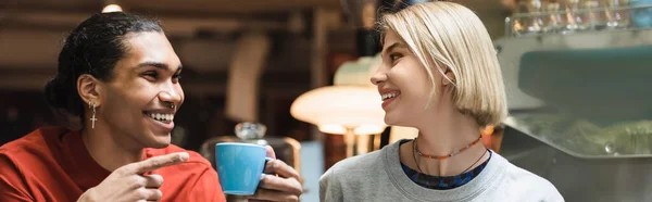 Lächelnder afrikanisch-amerikanischer Mann hält Tasse in der Hand und zeigt auf Freundin im Café, Banner — Stockfoto