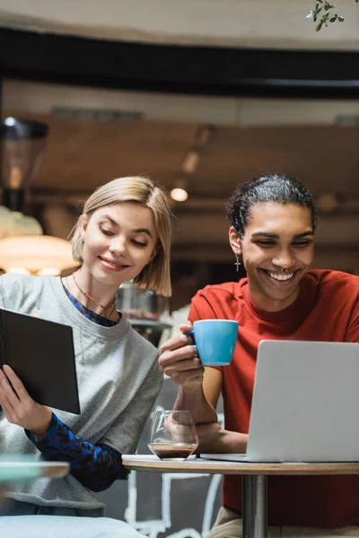 Des pigistes multiethniques souriants utilisant un ordinateur portable et un ordinateur portable près du café dans un café — Photo de stock