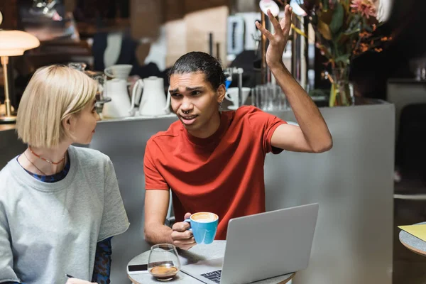 Freelancer afroamericano hablando con un amigo cerca del café y dispositivos en la cafetería - foto de stock