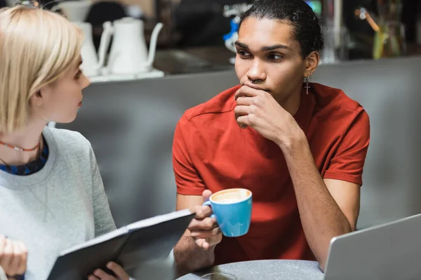 Jeunes pigistes multiethniques tenant du café et un ordinateur portable près d'un ordinateur portable dans un café — Photo de stock