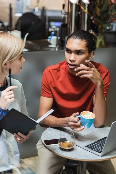 Freelancer afroamericano sosteniendo café y hablando con un amigo con portátil cerca de dispositivos en la cafetería - foto de stock