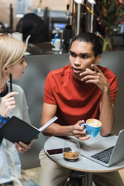 Libero professionista afroamericano che tiene il caffè e parla con un amico con notebook vicino a dispositivi in caffè — Foto stock