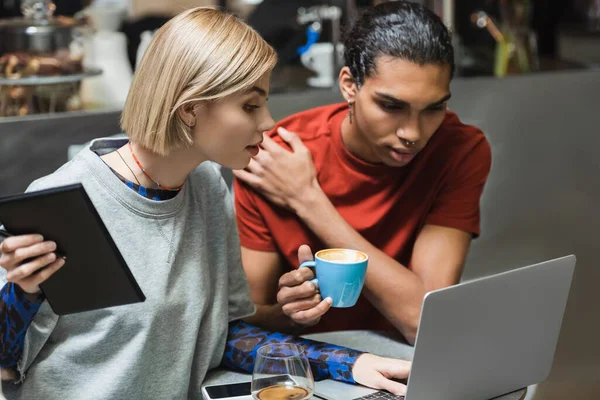 Joven freelancer con notebook usando laptop cerca de amigo afroamericano y café en cafetería - foto de stock