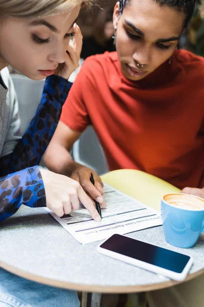 Young interracial friends looking at document near smartphone and coffee in cafe — Stock Photo