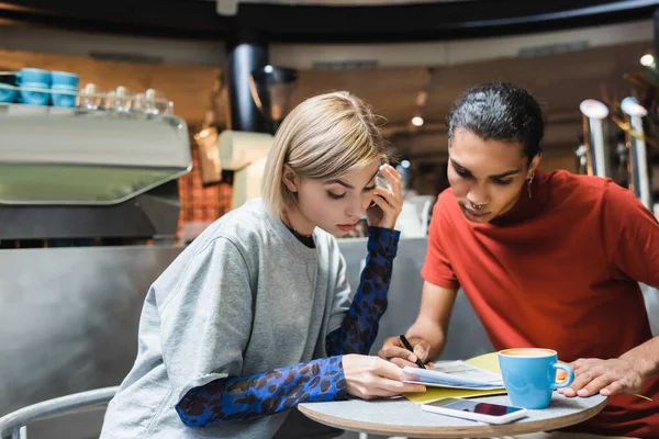 Amigos interraciales mirando los papeles cerca de teléfono inteligente y café en la cafetería - foto de stock