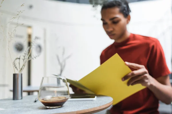 Caffè in vetro e smartphone vicino sfocato uomo africano americano in possesso di cartella di carta in caffè — Foto stock