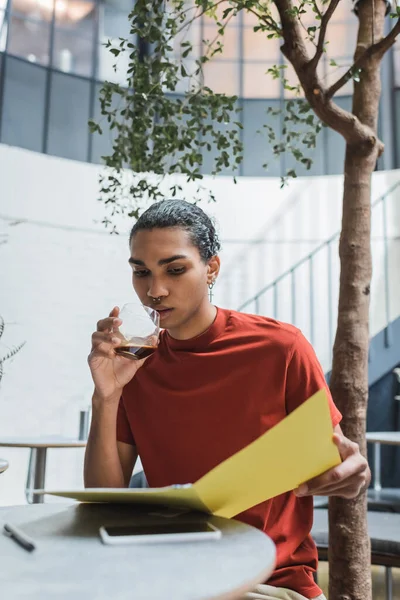 Hombre afroamericano joven sosteniendo un vaso de café y una carpeta de papel cerca de un teléfono inteligente en la cafetería - foto de stock