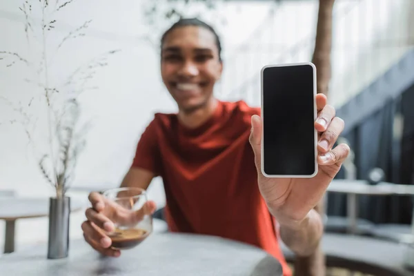 Smartphone con pantalla en blanco en la mano del hombre afroamericano borroso con café en la cafetería - foto de stock