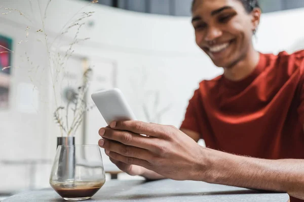 Uomo afroamericano offuscata utilizzando smartphone vicino vetro di caffè in caffè — Foto stock