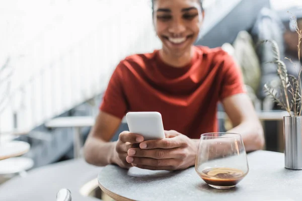 Cellulaire dans les mains d'un homme afro-américain flou près d'un verre de café dans un café — Photo de stock