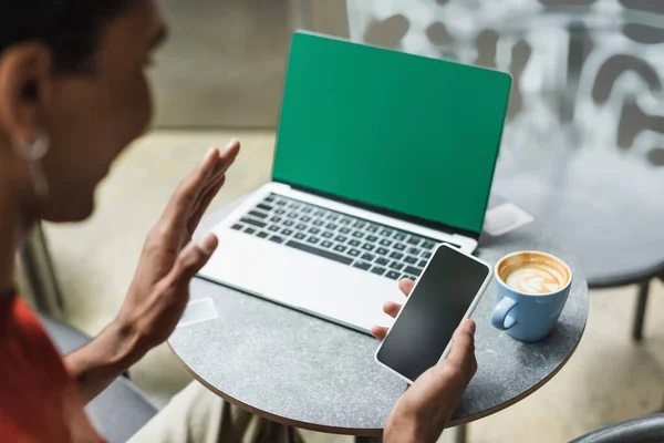 Desenfocado afroamericano freelancer tener videollamada en el teléfono inteligente cerca del ordenador portátil y café en la cafetería - foto de stock