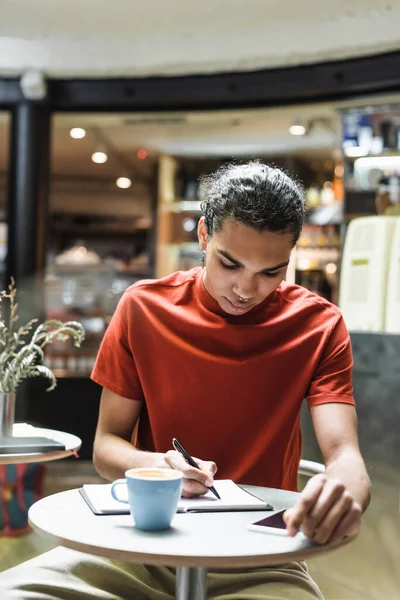 Uomo afroamericano che scrive su notebook vicino a caffè e smartphone nel caffè — Foto stock
