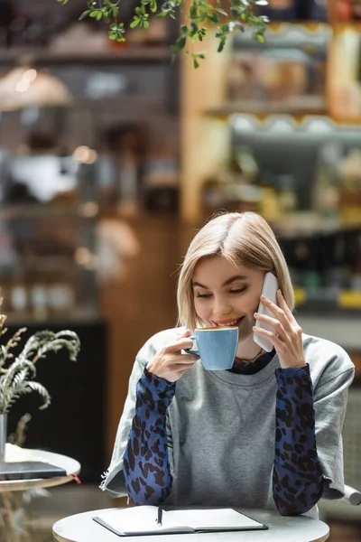 Mujer positiva hablando en el teléfono inteligente y beber café cerca de portátil en la cafetería - foto de stock