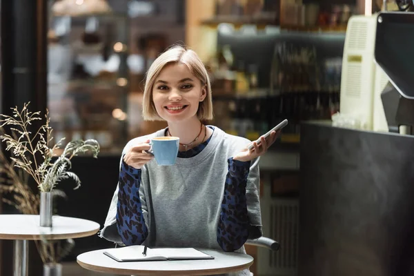 Femme blonde souriante tenant un téléphone portable et un café près d'un ordinateur portable dans un café — Photo de stock