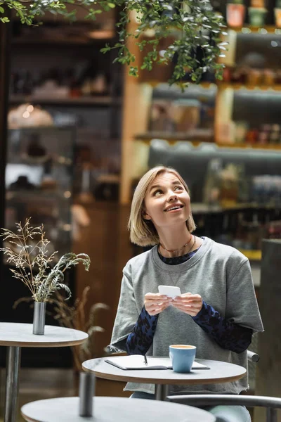 Mujer rubia sonriente usando teléfono inteligente y mirando hacia arriba cerca de café y portátil en la cafetería - foto de stock