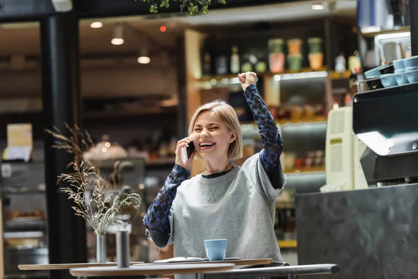 Mujer rubia emocionada hablando en el teléfono inteligente cerca de café y portátil en la cafetería - foto de stock