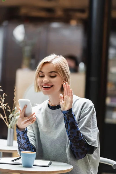 Femme positive ayant appel vidéo sur smartphone près de café et ordinateur portable dans le café — Photo de stock