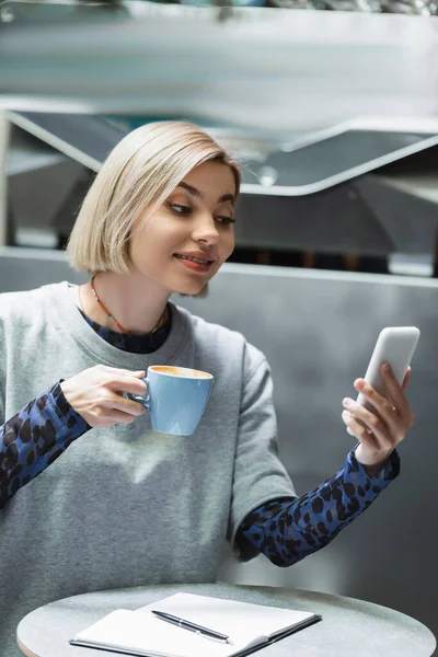 Mujer rubia joven sosteniendo la taza y el uso de teléfono inteligente cerca de portátil en la cafetería - foto de stock