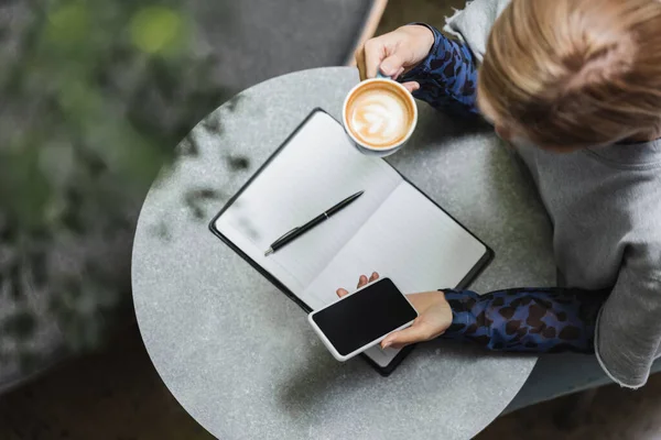 Overhead view of blonde woman using smartphone and holding cup of coffee near notebook in cafe — Stock Photo