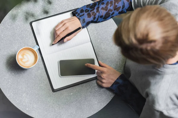 Vista aérea de la mujer utilizando el teléfono móvil cerca de café y portátil en la cafetería - foto de stock