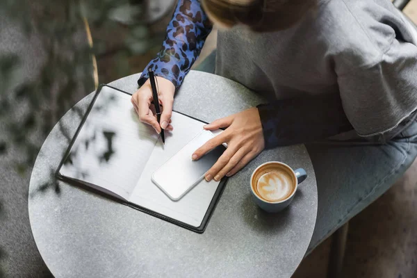 Visão geral da mulher escrevendo no notebook perto do smartphone e café no café — Fotografia de Stock