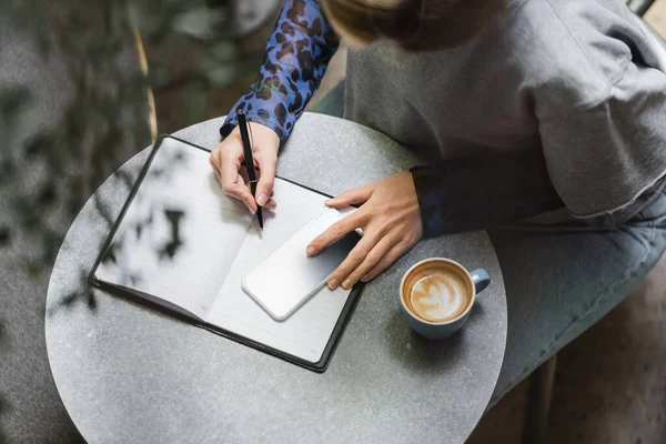 Overhead view of woman writing on notebook near smartphone and coffee in cafe — Stock Photo