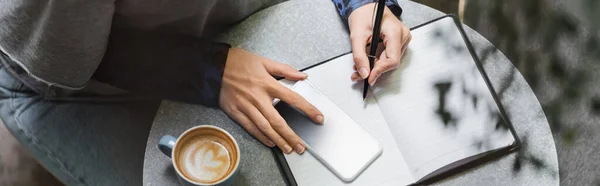 Vista cortada de mulher escrevendo no notebook perto de smartphone e xícara de café no café, banner — Fotografia de Stock