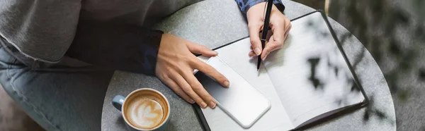 Cropped view of woman writing on notebook near smartphone and cup of coffee in cafe, banner — Stock Photo