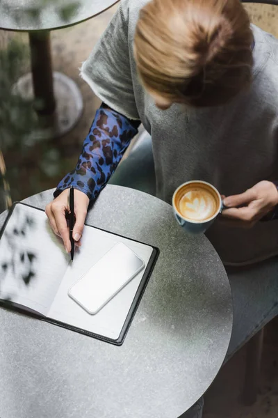 Visão aérea da mulher loira escrevendo no notebook perto do celular e segurando o copo no café — Fotografia de Stock