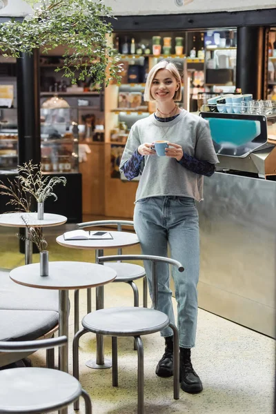 Mujer alegre sosteniendo taza de café cerca de teléfono inteligente y portátil en la cafetería - foto de stock