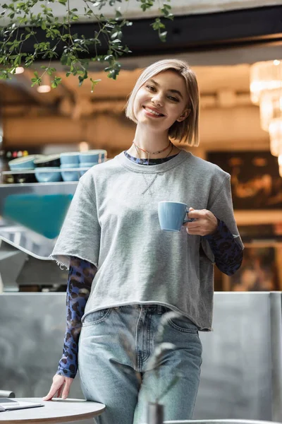 Mujer positiva sosteniendo la taza y mirando a la cámara en el café - foto de stock