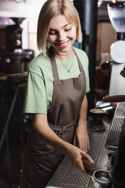 Sonriente rubia barista sosteniendo borrosa portafilter cerca de la máquina de café en la cafetería - foto de stock